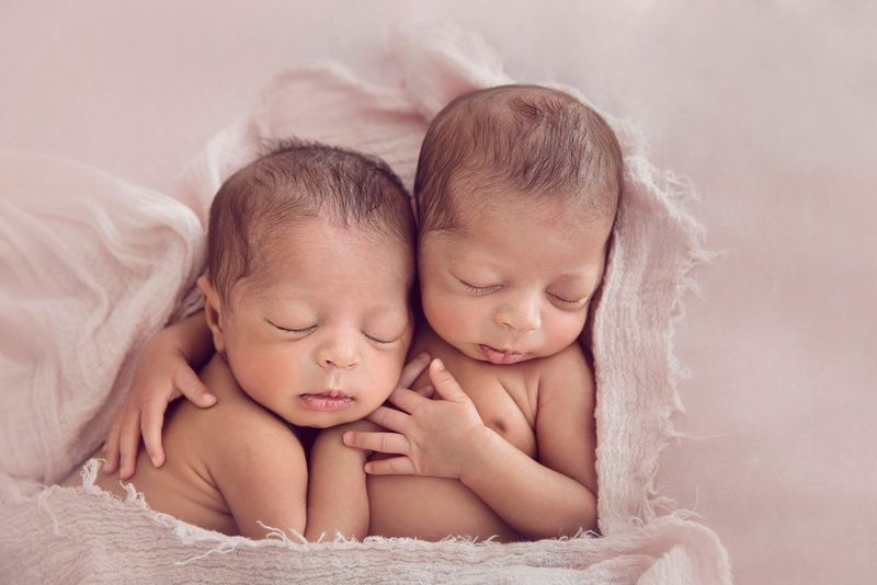 Два новорожденных близнеца, как две капли воды похожих друг на друга, спят рядом: один ребенок обнимает другого, завернутого в светлую ткань.