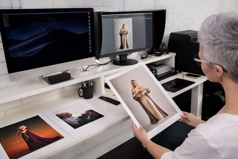 Человек, сидящий за белым столом с двумя мониторами и несколькими фотографиями на нем, держит изображение матери в развевающемся платье, качающей своего новорожденного ребенка.