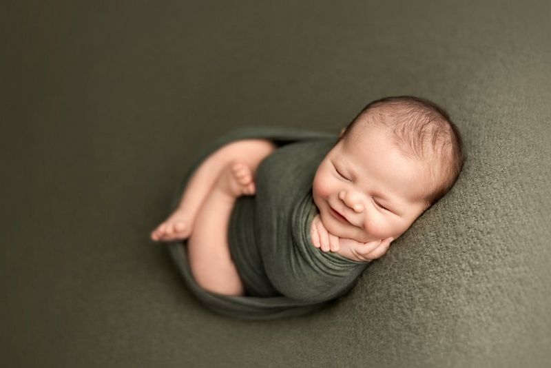 Крошечный младенец, завернутый в зеленую пеленку. Его ножки скрещены, он лежит щекой на одной руке и улыбается с закрытыми глазами.