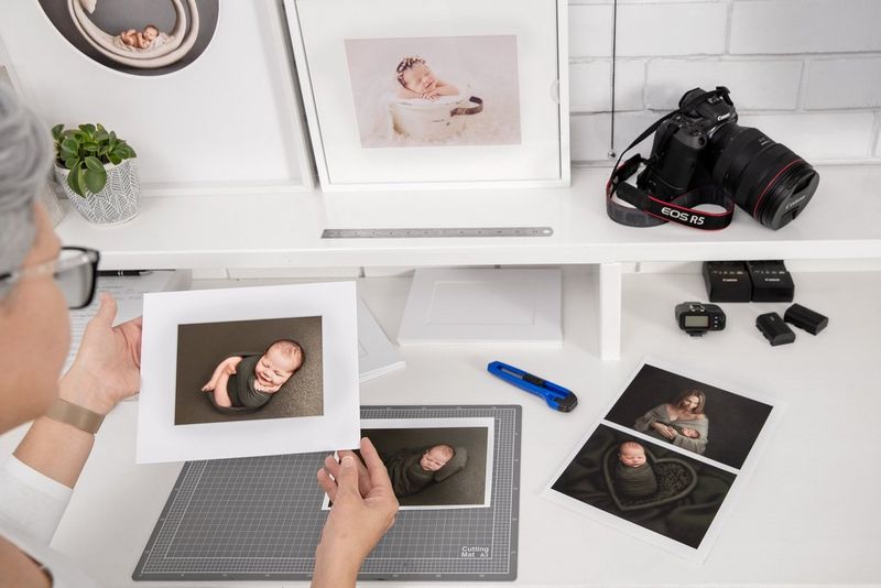 Человек сидит за белым столом, на котором лежит камера Canon и фотографии, и любуется снимком новорожденного ребенка в толстой белой рамке.