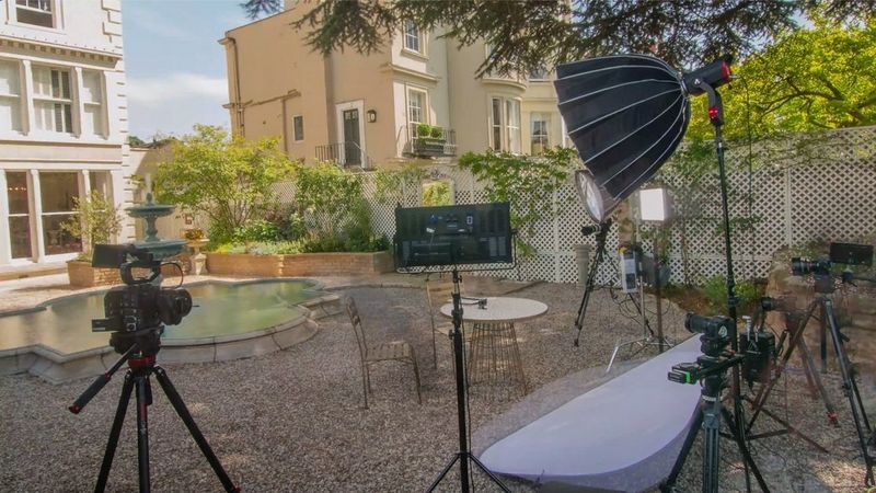 An array of Canon CInema EOS cameras on tripods and various kinds of lights surround a garden table and chairs outdoors.