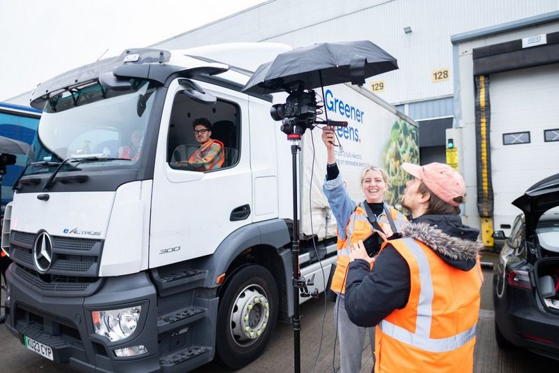  A crew member holds an umbrella over a Canon Cinema EOS camera mounted on a lighting stand, filming a lorry at cab height while the camera operator monitors an external display.