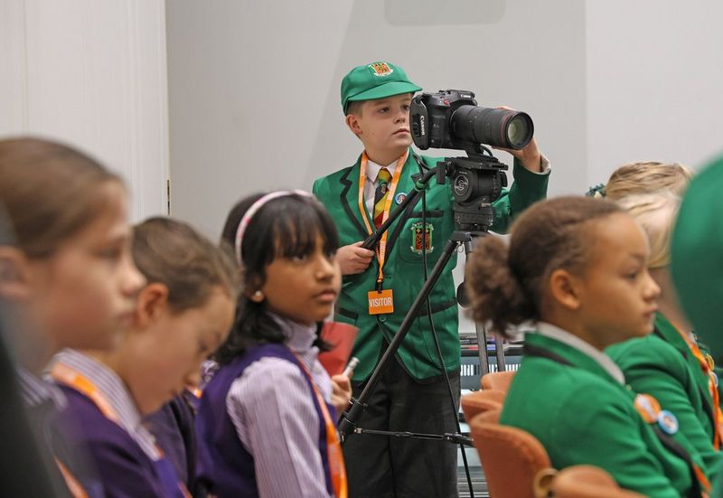 A child in a green school uniform operates a Canon Cinema EOS camera on a tripod behind other schoolchidren seated in rows.