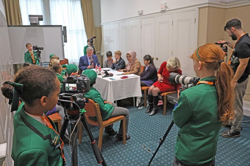 One adult technician and four schoolchidren operating Canon Cinema EOS cameras at each corner of a room. An audience of schoolchildren is watching people seated behind a white-draped table, taking part in a discussion.