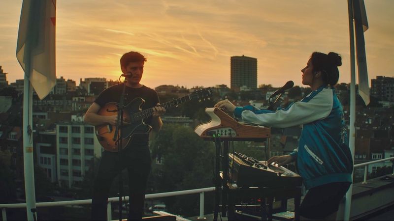На фоне заходящего солнца два музыканта стоят на крыше: один держит гитару, а другой играет на клавишах.