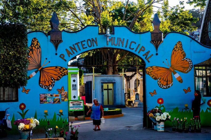 The entrance to a Mexican cemetery is painted bright blue, with large orange butterflies painted either side of the archway. A cleaner in a blue apron walks across the middle.