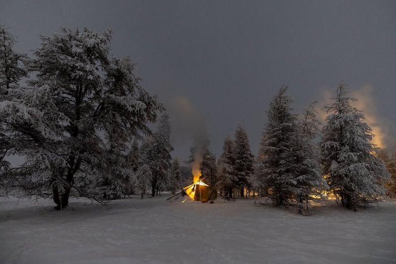 Een tent in een besneeuwd landschap, omgeven door bomen bedekt met sneeuw, terwijl een kampvuur achter de tent brandt, vastgelegd met een Canon EOS R3.