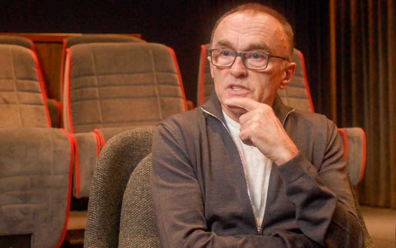 Regisseur Danny Boyle, met bril en één hand op zijn kin, denkt tijdens een interview in een kleine bioscoop na over een vraag.