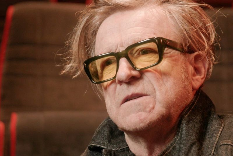 Close-up van de cinematograaf Anthony Dod Mantle met zwart-gele bril die wordt geïnterviewd in een kleine bioscoop.