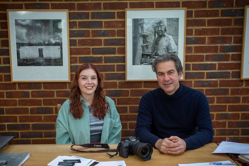 Due persone sorridono e siedono a un tavolo su cui è poggiata una fotocamera Canon EOS R6. Nel muro alle loro spalle ci sono diverse stampe incorniciate in bianco e nero.