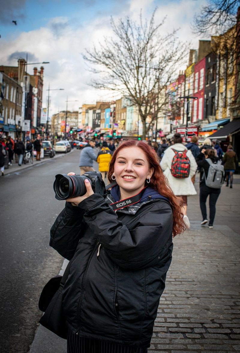 Una donna con lunghi capelli rossi e ricci in una trafficata via di Londra che regge una fotocamera Canon tra le mani sollevate.