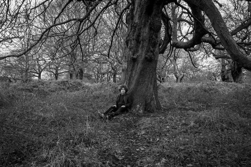 Un ritratto in bianco e nero di una figura seduta poggiata al tronco di un grande albero.