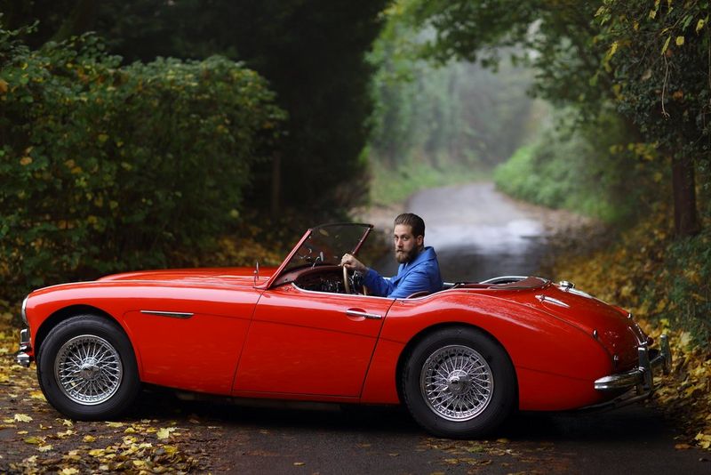 Un uomo indossa una maglia blu e siede al volante di una macchina sportiva rossa in un vialetto pieno di fogliame.