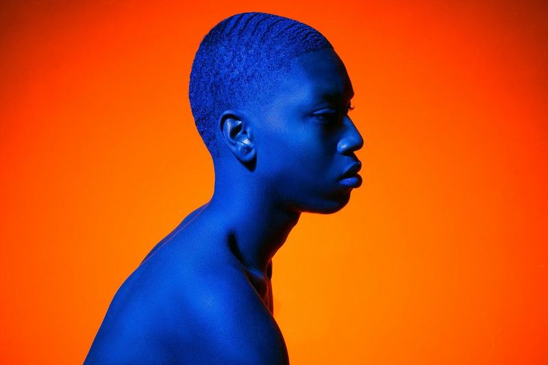 A head and shoulders shot of a topless man with shaved hair facing to the right. He is lit entirely in blue, against a warm orange background.