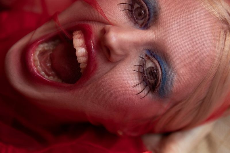 A close-up of a young woman with blonde hair, blue eyeshadow and elongated eyelashes. Her mouth is open as if she is about to scream. 