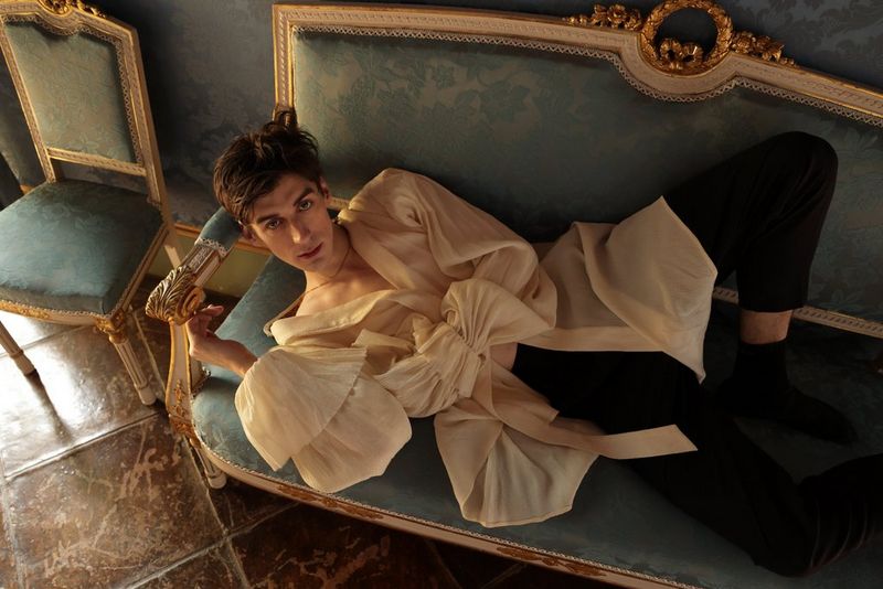 A man in a loose-fitting, floaty shirt lying on a green settee with a floral pattern and gold trim. 