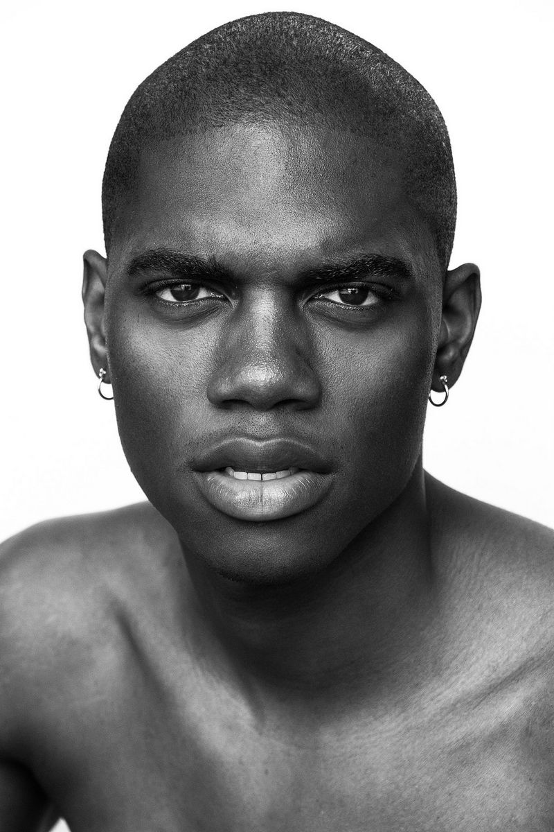 A young, bare-chested man wearing small hoop earrings stares intently at the camera. 