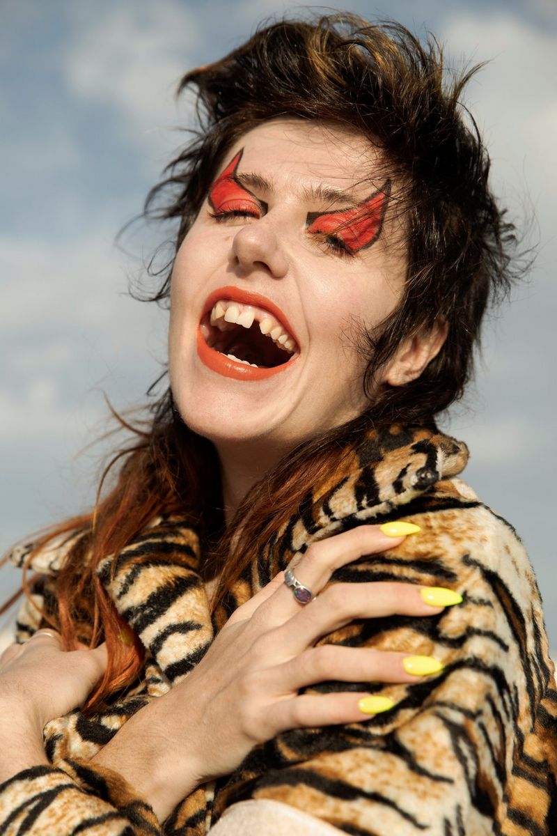 A woman in a fake tiger-skin fur coat crosses her arms across her chest and laughs. Her eyes are closed, showing devil horns painted across her eyelids and up onto her forehead.