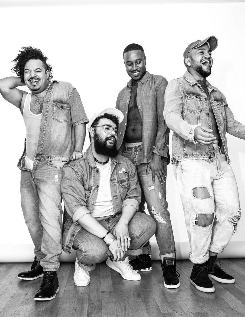 A black and white image of four men posing in denim clothing. They are laughing and smiling. 