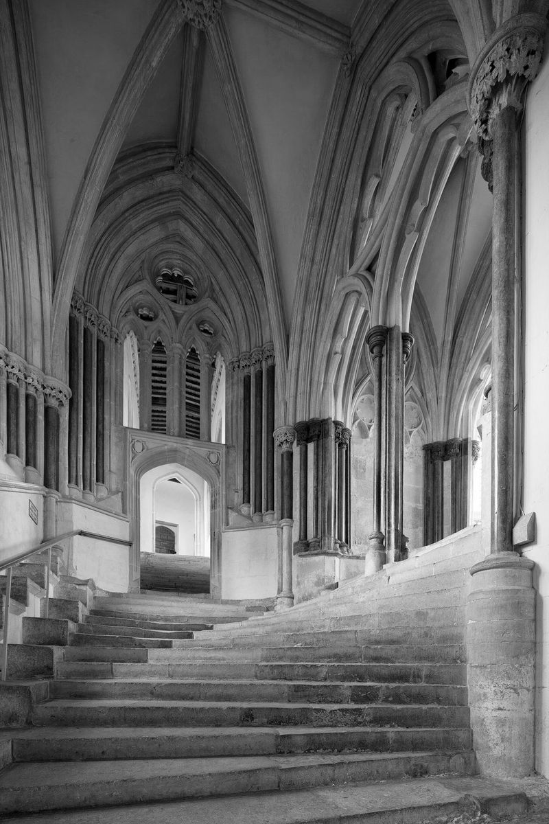 A black and white image of a winding stone staircase and ornate stonework inside Wells Cathedral.