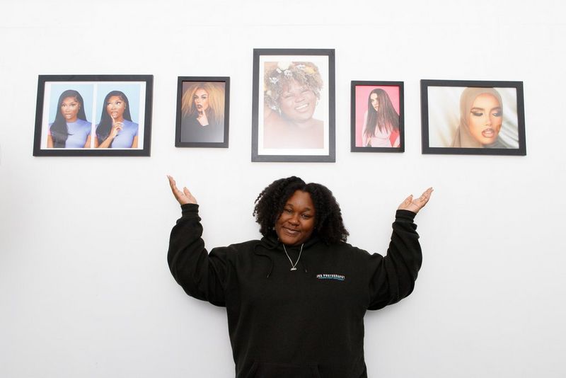 Fashion photographer Jade Keshia Gordon smiles at the camera and lifts her arms up to point at the five framed prints hanging on the white wall behind her. 