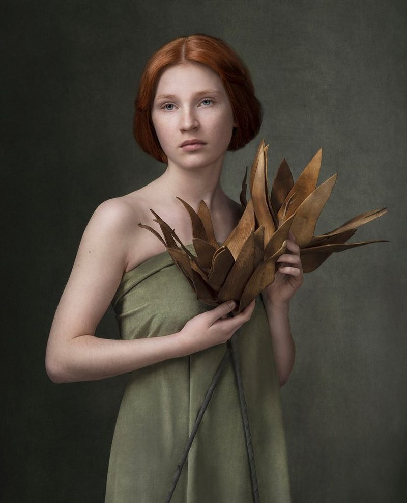 A fine art portrait of a red-haired woman holding a spray of foliage and wearing a light green dress matching the colour of her eyes, taken by Martina Wärenfeldt on a Canon EOS R5. 