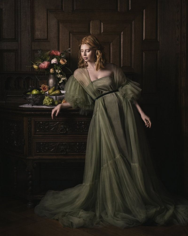 A fine art portrait of a woman in a flowing green dress resting her arm on a chest of drawers alongside an arrangement of flowers and fruit, taken by Martina Wärenfeldt on a Canon EOS R5. 