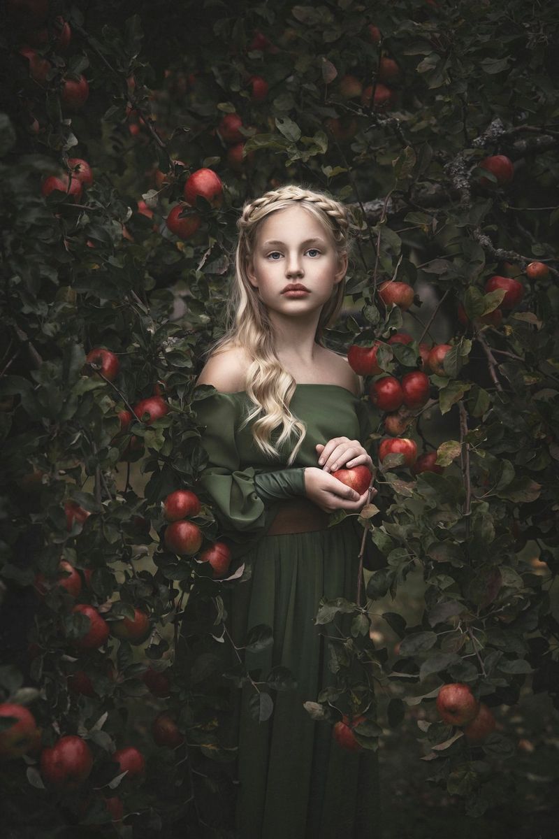 A fine art portrait of a young girl with long blonde hair and wearing a green dress standing in front of a tree laden with apples, taken by Martina Wärenfeldt on a Canon EOS R5. 
