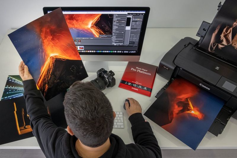 A man studies a portrait print of a photograph of Guatemala's Fuego stratovolcano. Next to him a Canon printer prints out a second image of the volcano, this time in landscape format. 