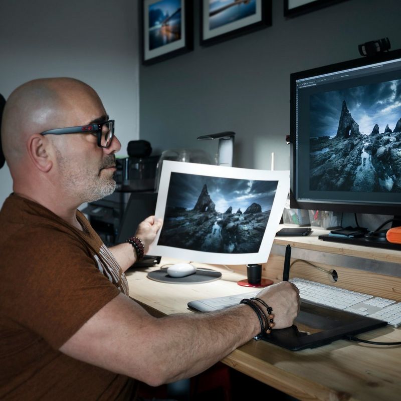 A person sits at a desk holding a print of a rocky landscape matching the one on the computer screen in front of them.