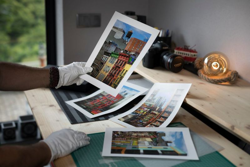 A white-gloved hand holds up a print of colourful buildings. Similar images can be seen on a nearby wooden desk.