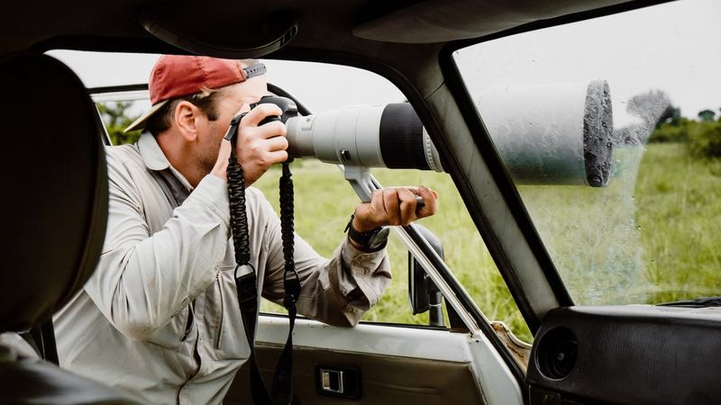 Фотограф выглядывает из окна транспортного средства в национальном парке Мерчисон Фолс в Уганде. Он держит в руках камеру Canon с большим телеобъективом.