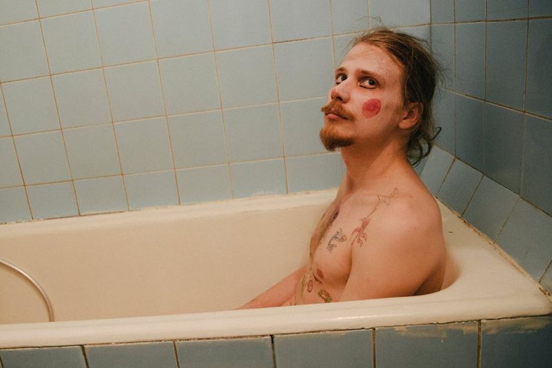 A topless man sitting in a bath wearing clown make-up.
