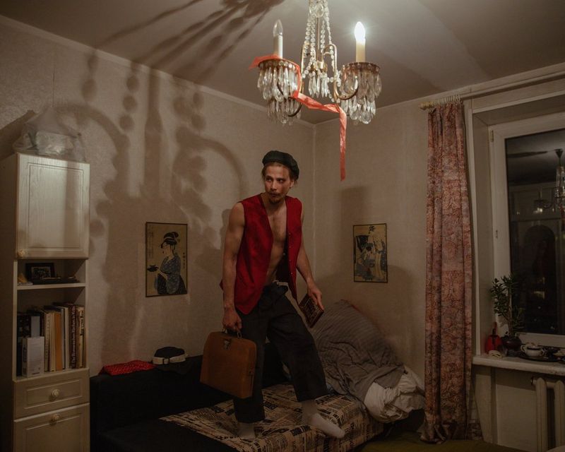 A man in a red waistcoat standing on a bed. A chandelier casts a shadow over the room. 