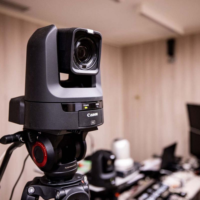 A Canon CR-N300 PTZ camera on a tripod, with a desk full of other AV equipment in the background.