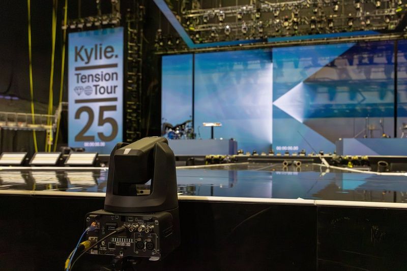 A rear view of the Canon CR-N700 at the end of a thrust stage in an empty arena, the camera pointing at large LED screens with "Kylie Tension Tour 25" displayed on one of them.