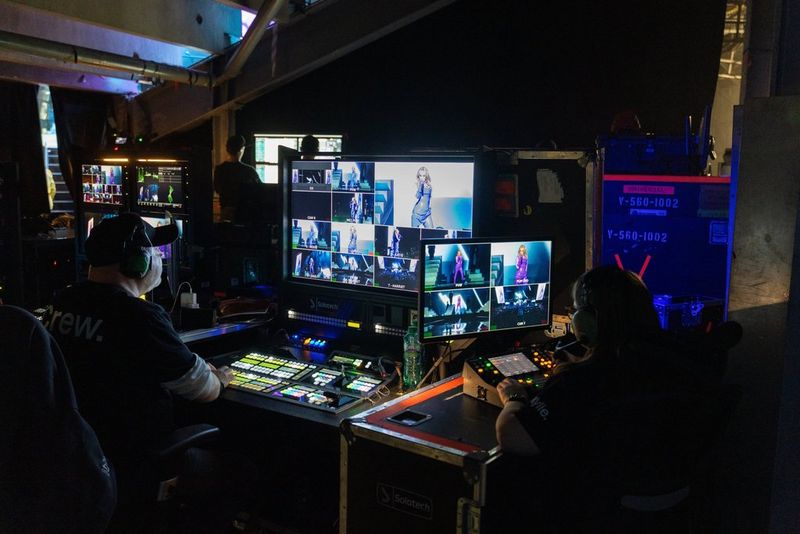Live video director Steve Price and PTZ camera operator Harriet Woollam sit in front of live monitors and controllers backstage in a dark area.