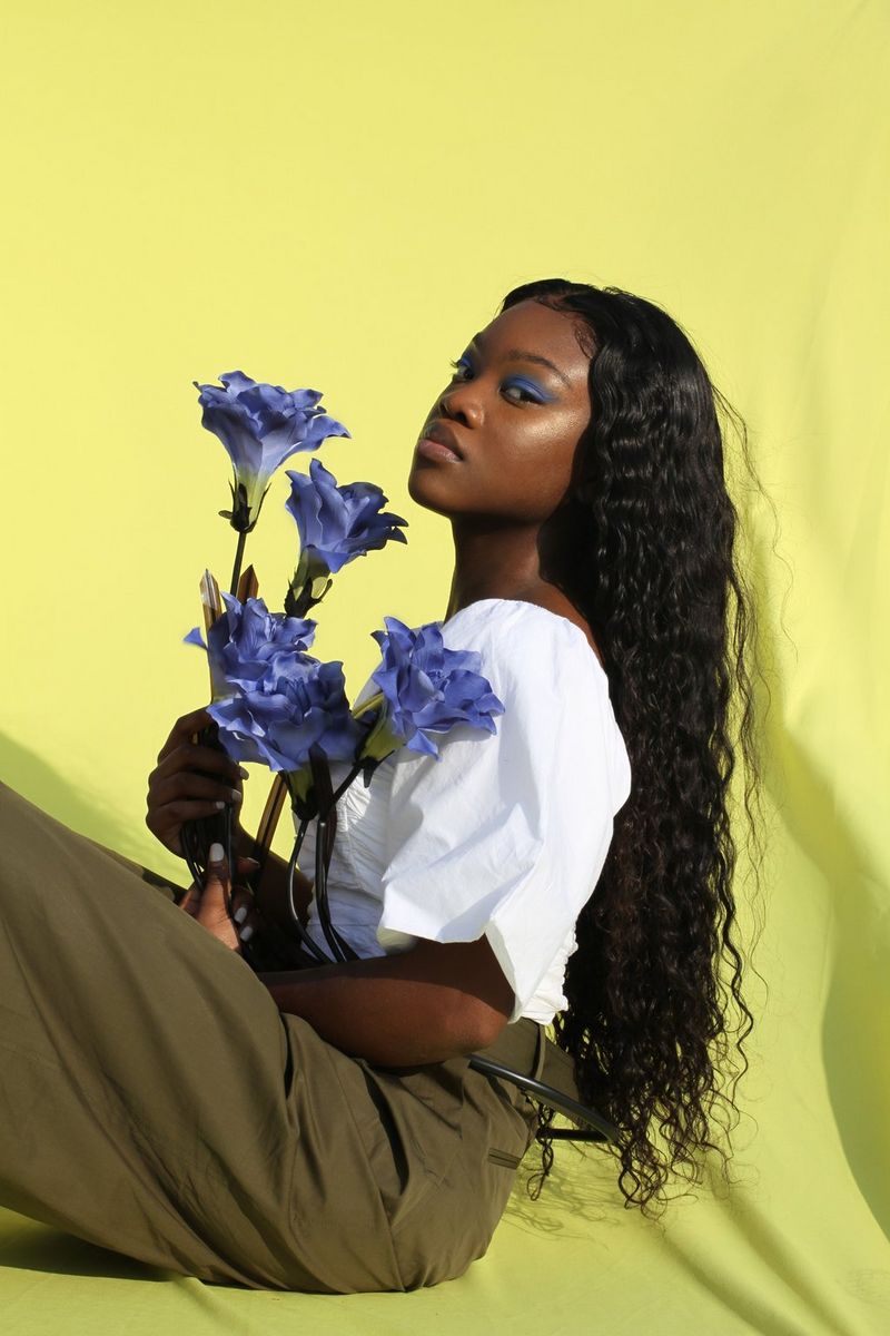 A young woman holding large purple flowers sits in front of a bright yellow background. 