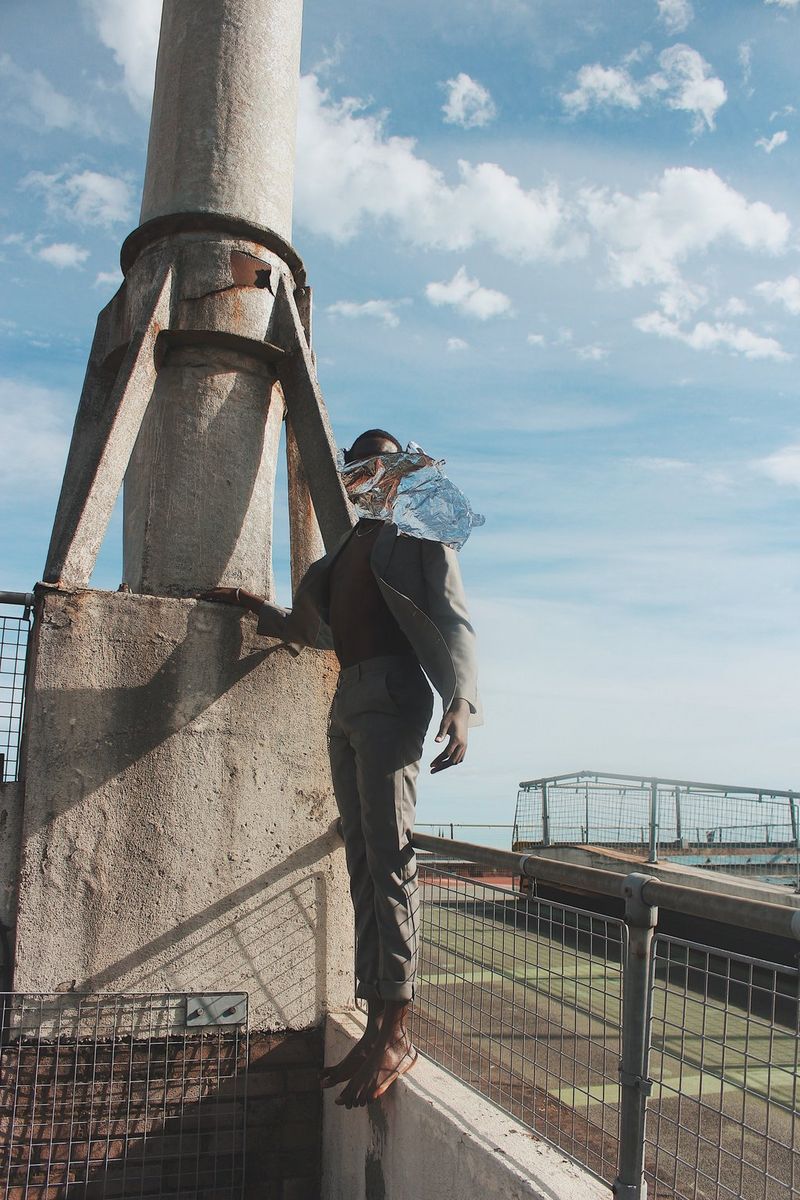 A man, his faced covered by silver foil, stands on the very edge of a wall next to a concrete chimney. 