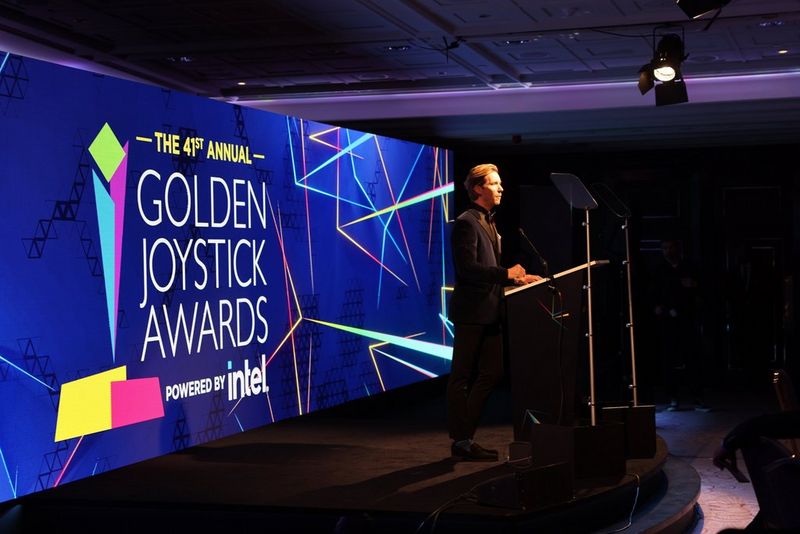 Ein Moderator steht auf der Bühne bei den 41. jährlichen Golden Joystick Awards. Die Bühnenkulisse beinhaltet ein grosses, farbiges Display mit geometrischen Designs und dem Logo der Veranstaltung.