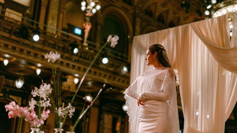Das Porträt einer Braut in einem großen Raum mit unscharfer Beleuchtung im Hintergrund und unscharfen Blumen im Vordergrund, aufgenommen mit einer Canon EOS R5 Mark II und einem Canon RF 50mm F1.4L VCM Objektiv von Hochzeitsfotografin Raïs De Weirdt.