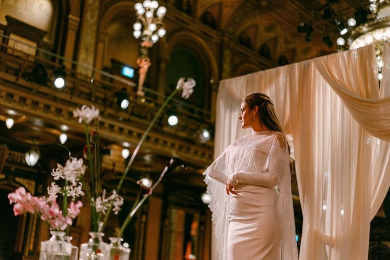 A low-angle portrait of a bride in a large room, with background lights and foreground flowers out of focus, taken on a Canon EOS R5 Mark II with a Canon RF 50mm F1.4L VCM lens by wedding photographer Raïs De Weirdt.