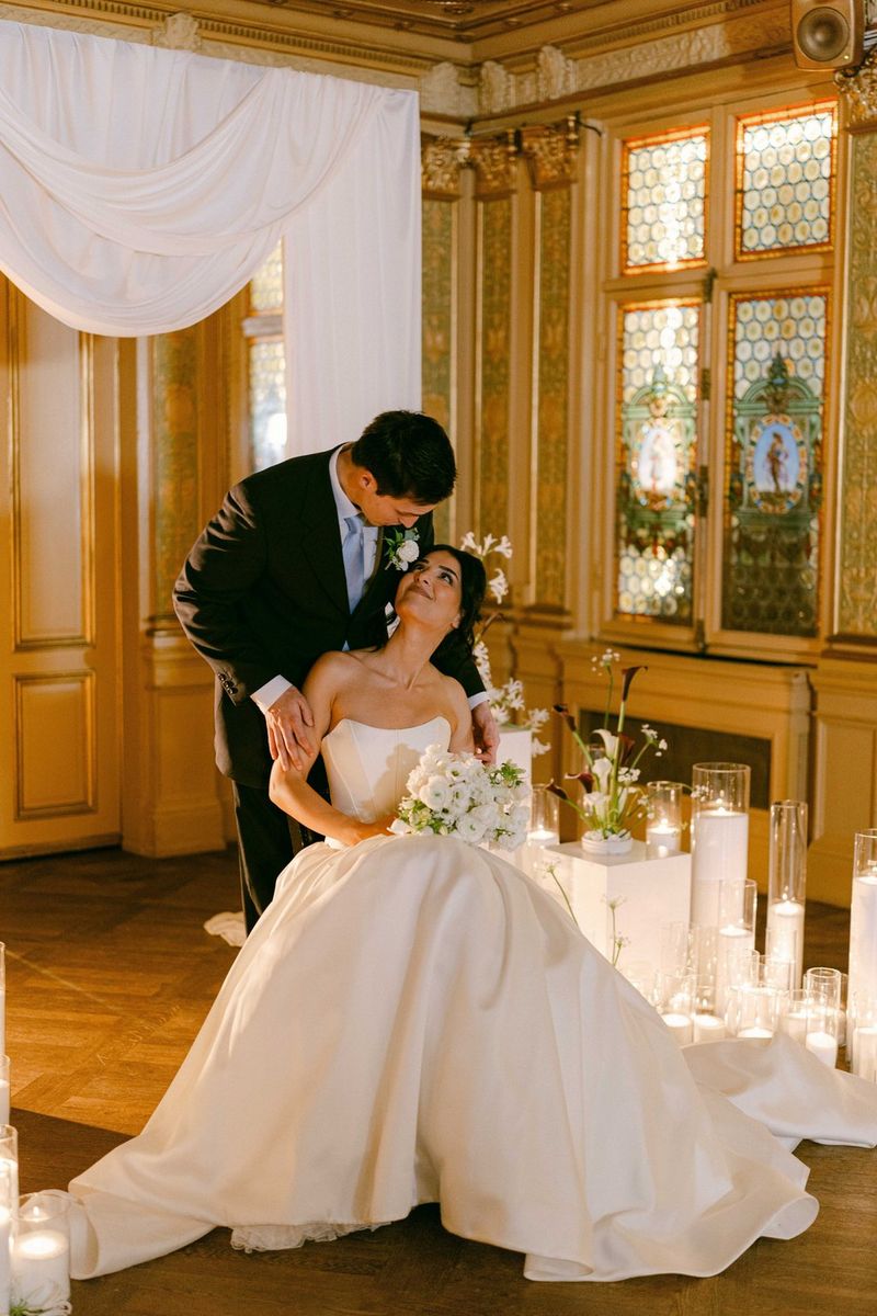 A wedding portrait of a bride seated and looking up at the groom leaning down to her, taken on a Canon EOS R5 Mark II with a Canon RF 50mm F1.4L VCM lens by wedding photographer Raïs De Weirdt.