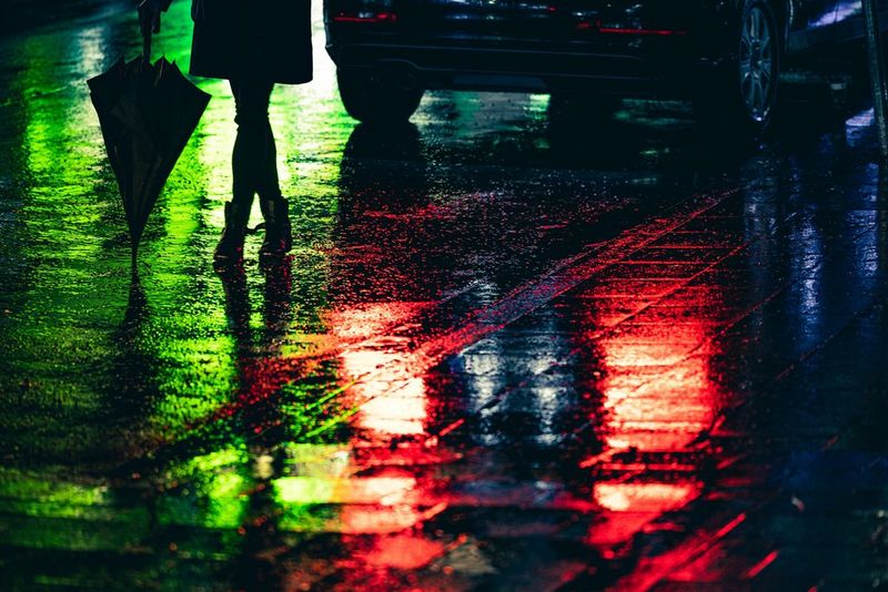 The bottom half of a woman silhouetted against a green neon reflection on the surface of a wet road. Red reflections from a car's brake lights are on the road beside her feet.
