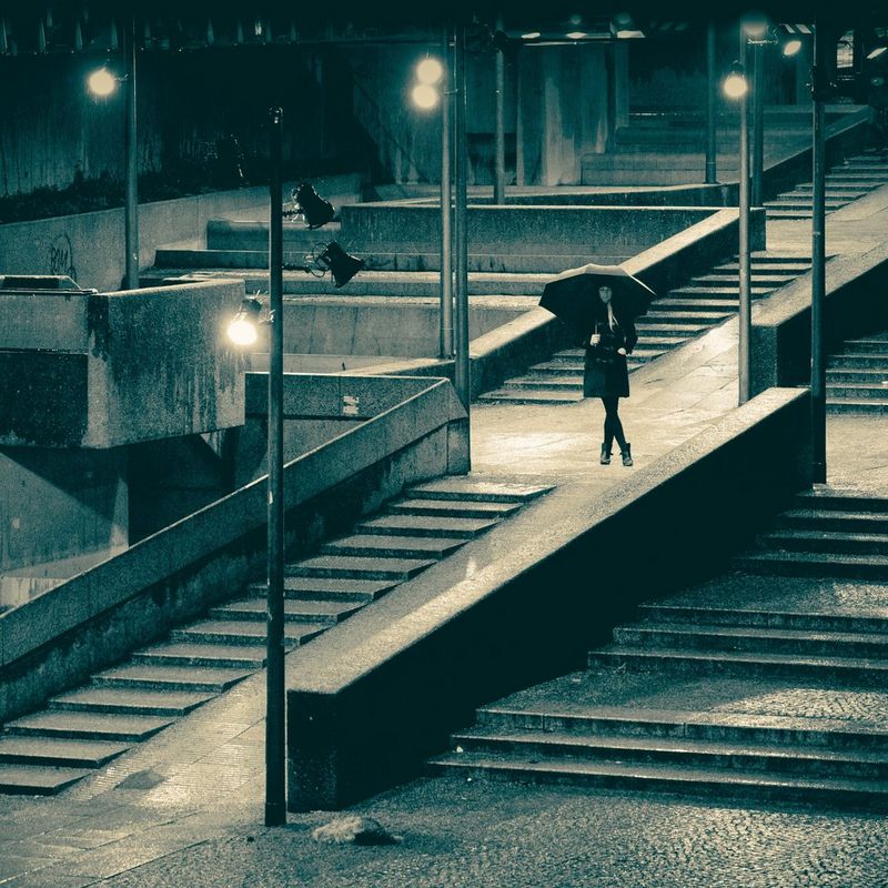 A woman stands holding an umbrella in the middle of a concrete flight of steps lit by streetlights.