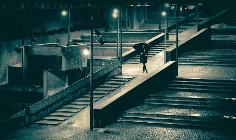 A woman stands holding an umbrella in the middle of a concrete flight of steps lit by streetlights.