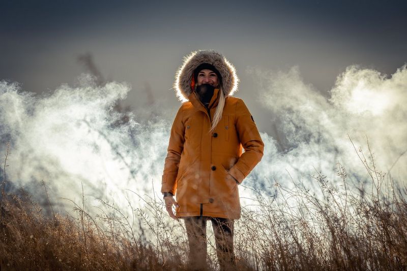 A woman in a yellow coat with a fur-lined hood stands in a field of long grass; smoke billows behind her.