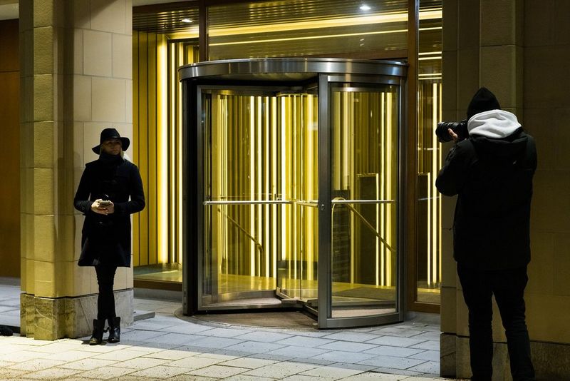 Action photographer Lorenz Holder with his back to the camera photographing a woman in a black hat and coat standing next to the entrance to a building.