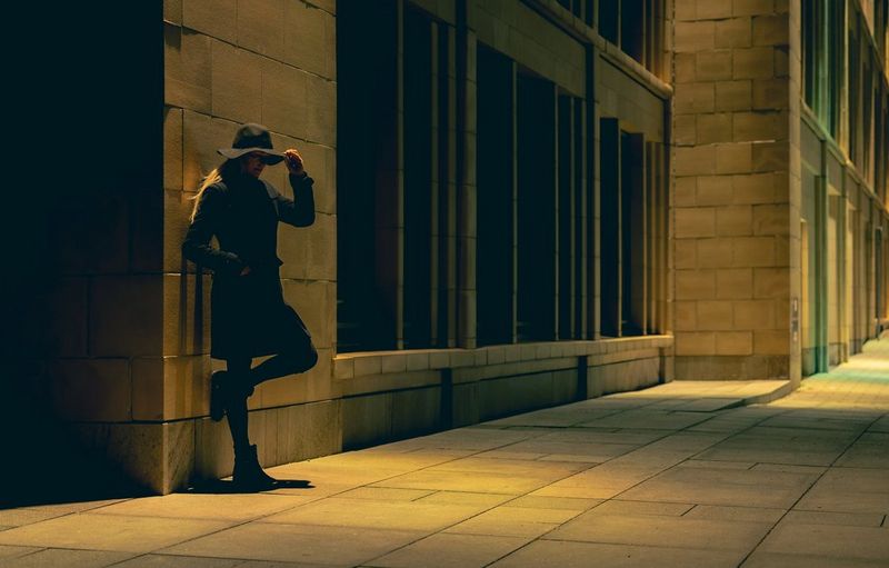 A woman leans against a building with one hand on her hat and one foot against the wall. The white balance settings have given the image an amber tone.