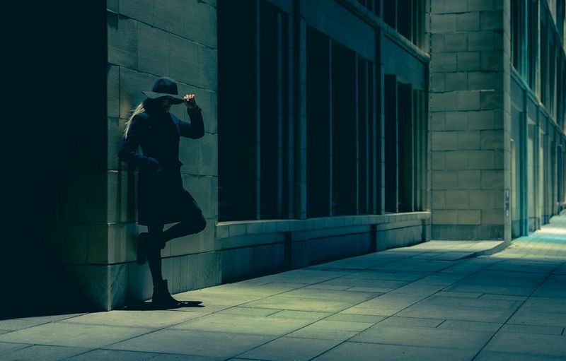 A woman leans against a building with one hand on her hat and one foot against the wall. The white balance settings have given the image a blue tone.